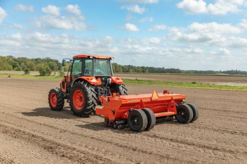 An orange tractor is driving through an empty dirt field to make precise rows for planting.